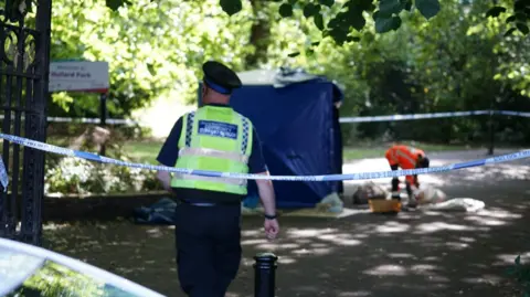 A blue and white cordon of police tape blocks off the entrance to a Manchester park. A uniformed Police Community Support Officer has his back to the camera, and in the background is a man in an orange hi-vis jacket standing beside a blue forensics tent, scooping matter into plastic bags. 