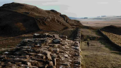 Clorroe Cam / The Spine Race A runner travels on a track next to Hadrian's Wall. It is afternoon based off the sunlight and there are small rocky bands to the left of the runner.