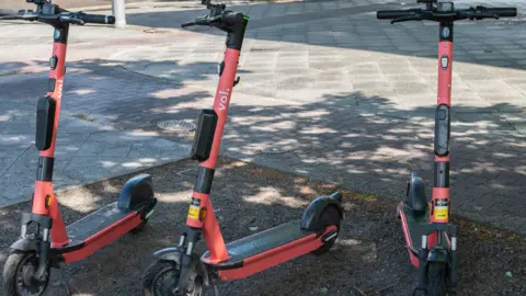 Stock image of Voi e-scooters parked at the side of a pavement. They have two wheels and are red with black handlebars. 