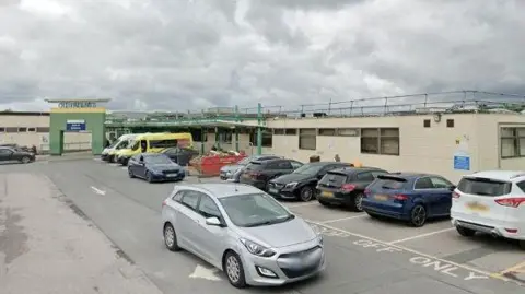 Google Maps The outpatient entrance and car park with several cars parked alongside the right side of the road