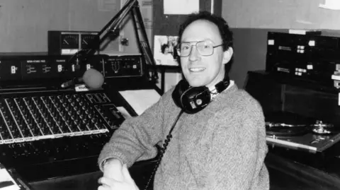 A black and white picture of a man with headphones around his neck wearing a jumper