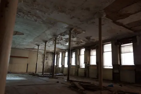 An empty room with tall columns and windows. The ceiling and floor show damage.