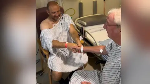 Manchester Royal Infirmary Ian Tonks (left) and Graham Sisson (right) in hospital gowns holding hands before surgery, seated beside a hospital bed 