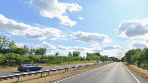 A dual carriageway with two cars driving away from the camera. The sky is blue with a few clouds.