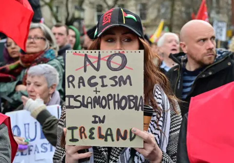 Pacemaker A woman holding a sign that reads No racism + Islamophobia in Ireland, she is wearing a black baseball cap with the Palestine flag on it