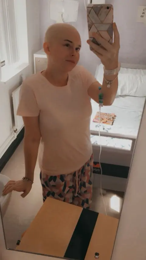 Fotografía tomada en el espejo de una habitación de hospital. En ella se ve a una persona con la cabeza rapada de pie junto a una cama, conectada a una vía intravenosa en un brazo. La persona lleva una camiseta de color claro y pantalones estampados, y sostiene un teléfono móvil frente a un espejo de pared.