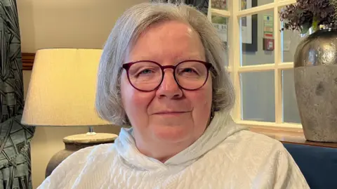 A woman with grey hair and glasses is wearing a white hoody and sat in an army chair near a lamp that is switched on