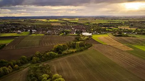 An aerial view of the Isle of Axeholme. There are several fields flanked by trees with buildings in the background. There are clouds in the sky lit up by the sun's rays.