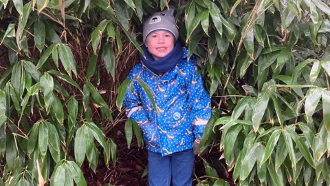 Evan standing under the cover of a large bush smiling at the camera. He is wearing blue jeans, a shiny blue and yellow jacket, a blue scarf and a grey bobble hat