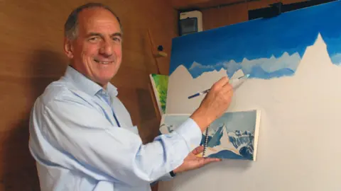 Kim Howells is holding a paintbrush to a canvas. He is wearing a blue shirt and appears to be in a wood-built building. The canvas has a snowy scene with white peaks in the foreground and bluer peaks in the background, giving a sense of depth.