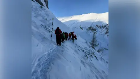 Salvamont Brașov A group of rescue personnel wearing red jackets, black backpacks and holding black walking poles are walking in a line along a very snowy hill with a steep drop below them