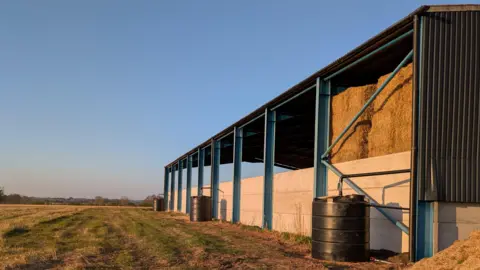 Shropshire Council The water butts at a farm. Three are lined up against a barn filled with hay. The photo was taken on a sunny evening with clear skies and trees in the distance.