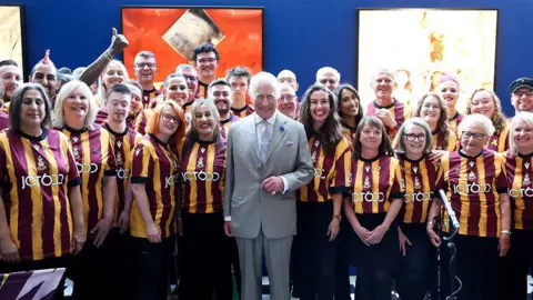 Getty Images King Charles in the centre of a crowd of people who were members of the Bantam of the Opera choir in Bradford. He is dressed in a grey suit and is smiling; members of the choir are dressed in Bradford City home shirts, which are amber and maroon.