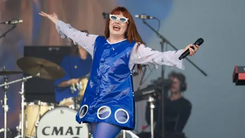 Sam Fender Belfast: Thousands heading to Belfast Vital gig 5 Samir Hussein/Getty Images CMAT performs on the Pyramid stage during the Glastonbury festival in June 2025. She is holding a microphone and has her arms outstretched. CMAT has long, straight auburn hair and is weaing a white-rimmed sunglasses, a white blouse adorned with silver Euro signs, a royal blue tunic made of a shiny patent material and bright blue tights.
