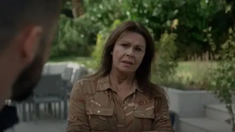 BBC/Left Bank Pictures Julie Graham, wearing a brown buttoned shirt, appears in front of the spacious green garden which has grey seating and table to the left.