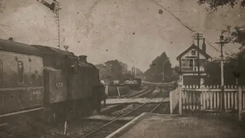 Edwin Williams A grey grainy photo of New Longton and Hutton railway station in 1963 showing a train on the tracks to the left and the signal box building to the right behind a white picket fence