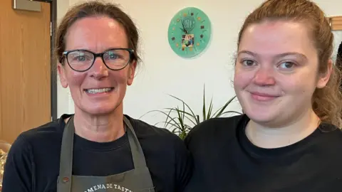 Jo Beattie with brown hair tied back, wearing black-rimmed glasses, a black top and a green overall, stands next to her daughter Nadia, who has light brown hair tied back and is also wearing a black t-shirt. 
