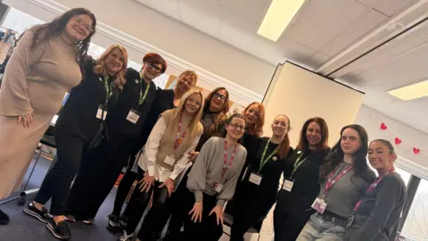 Kerry Galliano A group of 12 women standing together arm in arm smiling at the camera. They wear pink or green lanyards.