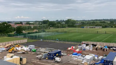 A construction site with containers, excavators and barriers. In the background is a row of sports pitches.