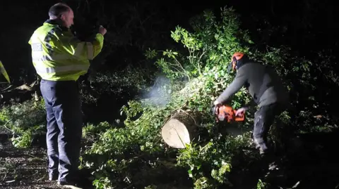 Northamptonshire Police Two people by a fallen tree. A police officer, to the left, is wearing a high-vis jacket and dark trousers. A man, in dark clothing and wearing an orange hard hat, holding a chainsaw is cutting a tree. There is lots of foliage around the tree. 