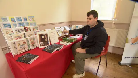 A man who is blind sits at a table full of braille versions books and cards that he has created. He wears a black hoodie and green trousers.