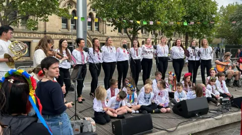 A line of women all wearing white singing outside the City Hall in Bradford. Children are sat in front of them and there are people playing guitars to the right of the image, and a person playing a violin and person playing a brass instrument to the left of the image.