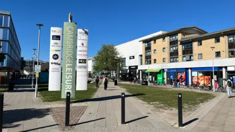 Cambridge Leisure Park, with a path and grassed area through the centre of it. To the left is the edge of a hotel. To the right are shops and a cinema. A big sign saying "Cambridge Leisure" in the foreground shows the different facilities based at the site. There are some people walking around and many bikes are parked near the shops.