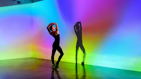 a female dancer in a black top and leggings holds a pose while her shadow is case on a multi-coloured background on a stage