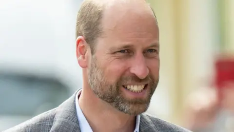 Prince William, the Prince of Wales, smiles as he is photographed outside. He is wearing a light-coloured casual jacket and a pale shirt with an open neck.