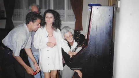 Norman Taylor A photo of a young woman with dark curly hair playfully grimacing next to an arcade machine in a pub. A man grins next to her as he leans over towards the machine, while an older woman with grey hair bends towards the machine behind them.