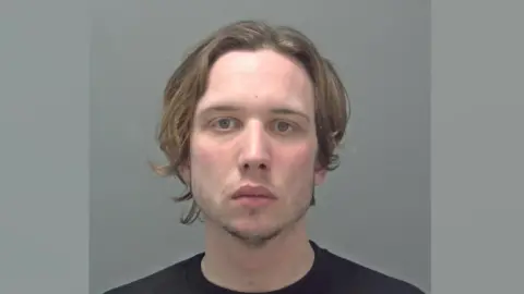 A mugshot head and shoulders image of Elliott Mallett. He is wearing a black t-shirt and standing against a grey wall. He is staring into the camera.