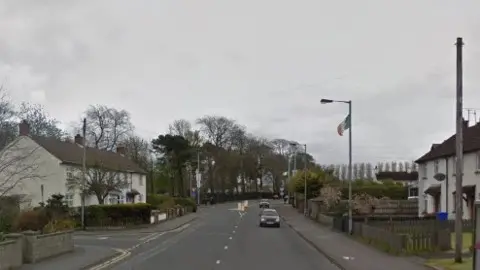 A street in Limavady can be seen with two cars driving in the same direction. There are houses and lampposts of either side of the footpath. A lamppost on the right has an Irish flag flying from it. 