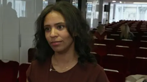 Lauren stands in the foreground of what appears to be a lecture hall, conference room, or auditorium. She is positioned slightly to the right of the centre of the frame and is facing toward the left side of the image. She has shoulder-length dark wavy hair and is wearing a dark reddish-brown top along with a thin necklace.