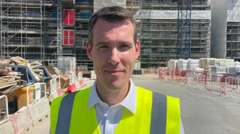 Housing and Planning Minister Matthew Pennycook smiles facing the camera. He is standing in a building site and wearing a yellow hi-viz vest.