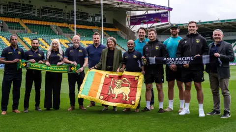 Northampton Saints A group of men and women pose for a picture. Among them is Julia Chapman, dressed in a green blazer. A Northampton Saints scarf and an Asia Pacific Lions are being held up along with a Sri Lankan flag.