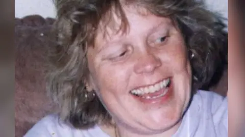 Debbie Griggs looks off camera and is smiling. She is wearing a pale coloured t-shirt and has short light brown hair.