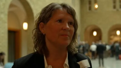 Merete in a large hallway. She has shoulder length wavy brown hair and is wearing a white shite and dark suit jacket