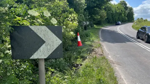 A black sign with an arrow on it on the side of the A36 in Wiltshire. The road next to the sign curves to the right. There are two cars travelling in opposite directions on the road. There is a traffic cone on the grass verge near the sign.