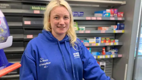 A woman in a blue top with blonde hair stands in front of shelves in a store