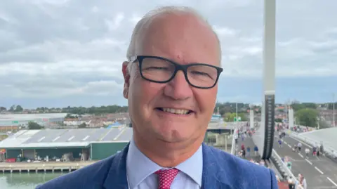 Matthew Hicks is standing on the Gull Wing Bridge. He is wearing a blue suit, blue shirt and a pink patterned tie. He is wearing black-framed glasses. Behind him to the left, a river is partially visible, and to his right behind him is a road bridge. It is cloudy above.