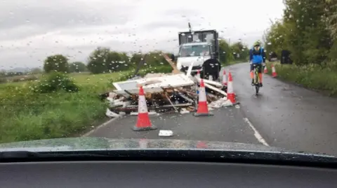 Lee Colvin A large pile of what appears to be building waste is dumped across a narrow, rural road. It has been cordoned off, as one cyclists rides past it.