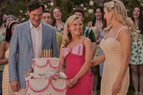 Kimberley French/Prime Video A man and two women smiling at each other, standing infront of a two-tierd cake, with gold candles and pink decorations. People are in the background, also smiling.