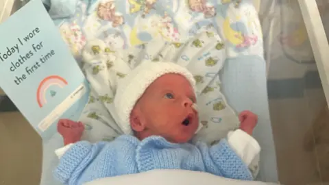 Family handout Jude in the cot in the neonatal unit. He is holding a card saying 'Today I wore clothes for the first time'. He is wearing a blue knitted cardigan and a white knitted hat. He is looking to the side of the cot, with his mouth open in a surprised or excited espression.