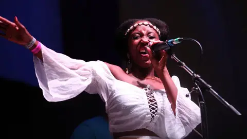 Getty Images Nadine Caesar of Soul II Soul performs during Weyfest Festival 2021wearing a white shirt and singing in to a microphone with one hand in the air