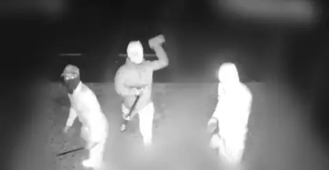 Avon and Somerset Police CCTV image shows three people, with their faces covered, carrying weapons and looking into the camera. One person had his arm above his head about to throw a brick. 