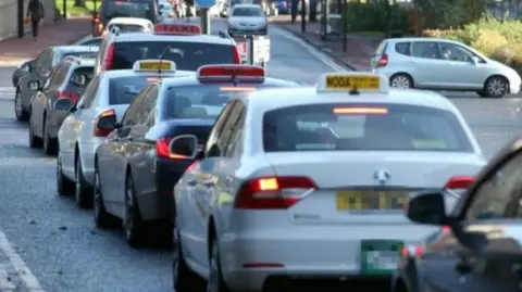 A row of private hire cars and taxis on a street, with signs on their roofs