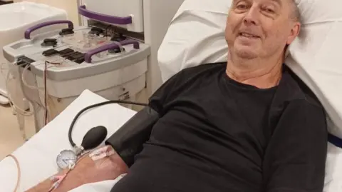 Steve Cervi Steve Cervi, who has a bald head and is clean shaven, lies on a hospital bed wearing a black t-shirt. A cannula is attached to his right arm with medical tape and medical machinery is visible in the back ground. 