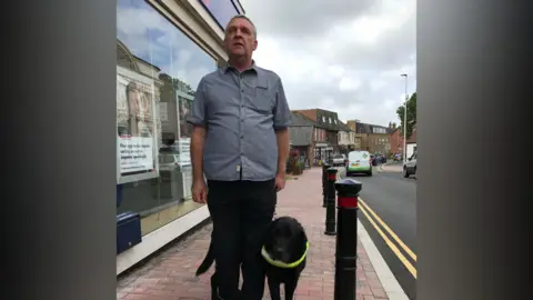 A man, Steve Saunders, stood by a road with a guide dog.