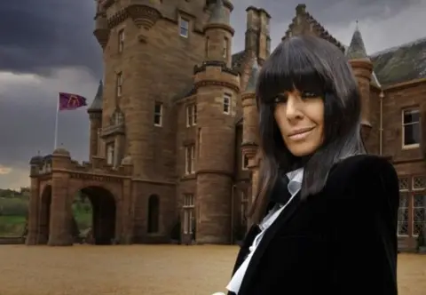 Claudia Winkleman standing outside the Traitors castle, with dark cloudy sky in the background