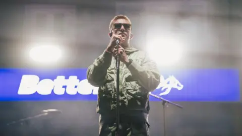 Luke Brennan/Getty Images Jonny Yerrell of The Reytons is wearing a green parka jacket and is holding a microphone. 